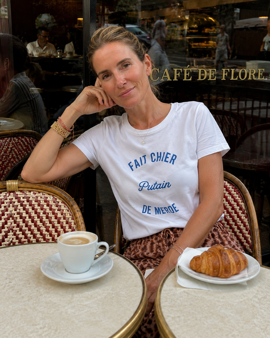 Woman sitting at a cafe table with a coffee and croissant, wearing a white t-shirt with blue text.