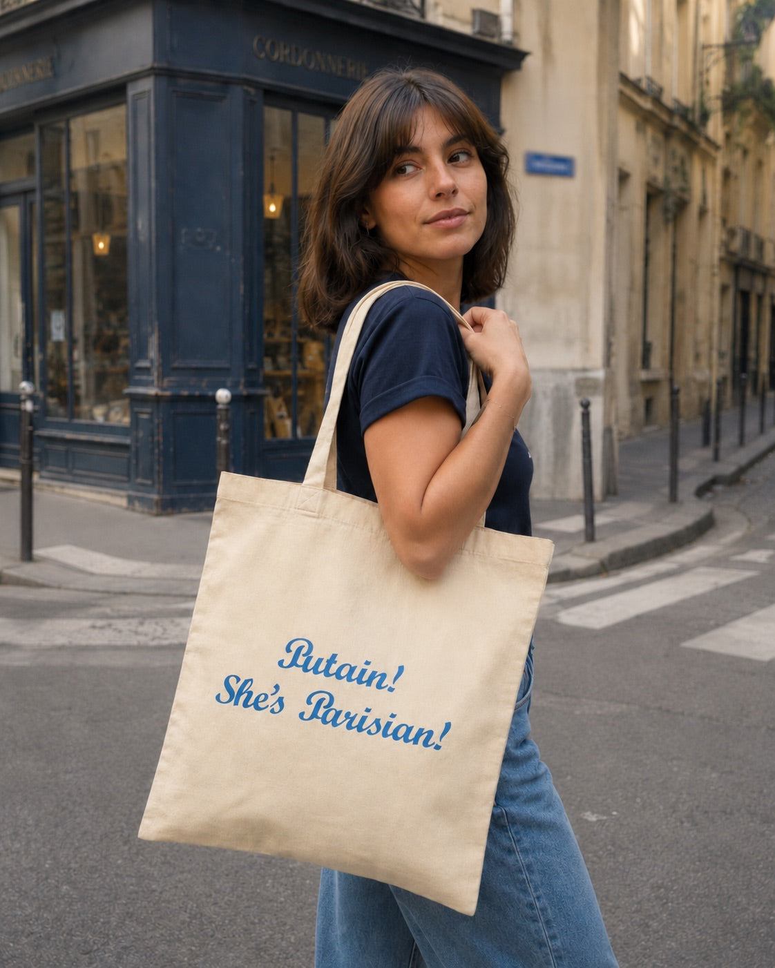 Woman holding a tote bag with 'Putain! She's Parisian!' text on a city street.
